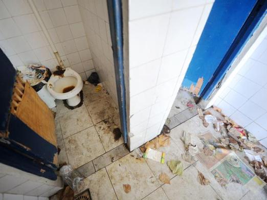 O banheiro ao lado da farmácia tem fezes espalhadas pelo vaso sanitário e sujeira acumulada em toda a parte